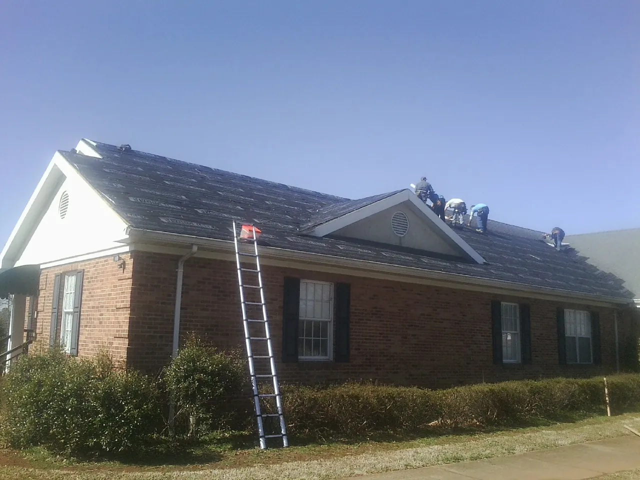 Crew tearing shingles off a brick ranch roof with a ladder set — Emergency Roof Repair work by our crew in Newland