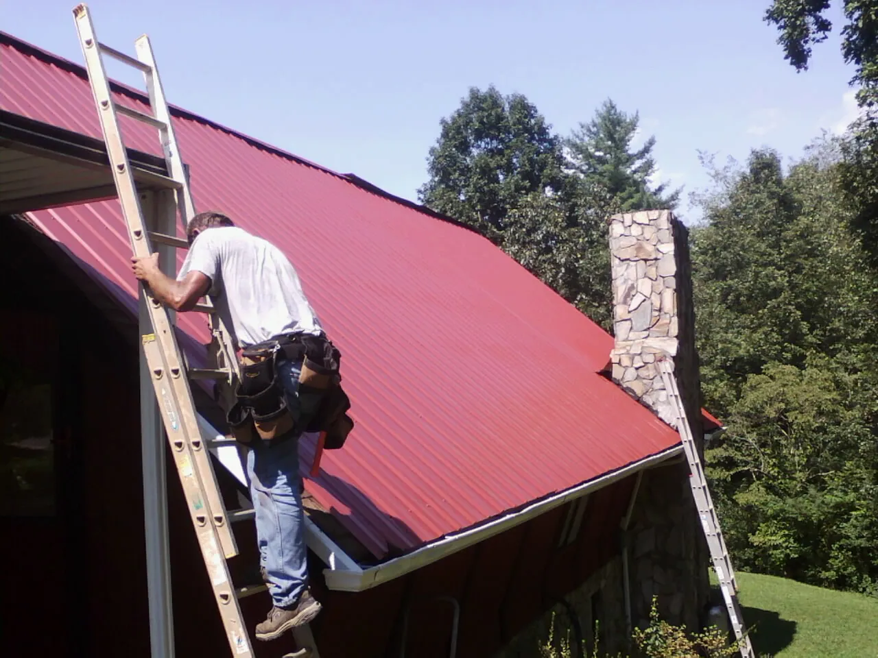 Roofer on a ladder beside a red metal roof and stone chimney — Soffit & Fascia Repair by Roofing Brothers in Newland